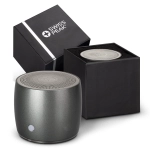 A round grey speaker with a textured top next to its black packaging, featuring a logo on the box.