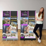 A pull-up banner featuring a real estate promotion with a woman standing beside it. The banner has purple, black, and white colours.