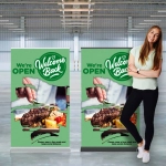 Two green pull-up banners featuring a dish of food and a welcoming message, displayed in a spacious venue with a woman beside them.