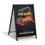 A black A-frame sign featuring a promotional graphic with gold text and a ribbon design for a grand opening event.