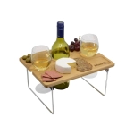 A wooden picnic table with two glasses of white wine, a bottle, cheese, crackers, olives, and grapes.