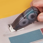 A grey and black glue tape dispenser with a white base being used on paper. It has a logo on it.