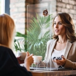 A clear personal protection screen is set between two people seated at a table, with plants in the background.