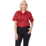 A woman models a red button-up short sleeve shirt paired with dark jeans, featuring a logo on the shirt.