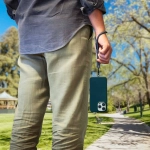 A person holding a dark green phone secured by a black and white patterned eco wrist strap, standing in a park.