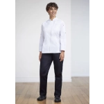 A woman stands wearing a white uniform top and navy scrub bottoms, paired with black shoes, in a bright indoor setting.