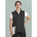 A woman wearing a black vest with a zip front, holding a clipboard, against a light green background.