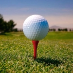 A red golf tee holding a white golf ball on a grassy surface, with a blue sky in the background.