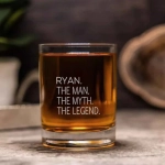 A glass tumbler with a clear body holds a drink, featuring a logo and an engraved phrase.