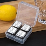A transparent container holding four stainless steel whiskey ice cubes, alongside a lemon and a glass on a wooden surface.