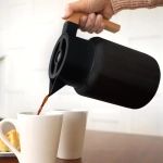 A drink bottle in matte black with a wooden handle, pouring liquid into two white cups on a wooden table.