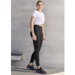 A woman wearing black flex leggings and a white tied T-shirt, standing against a textured wall. The leggings have a logo.