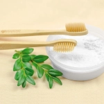 A bamboo toothbrush with soft bristles is placed next to a bowl of white powder, accompanied by green leaves.