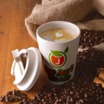 A reusable coffee cup in light beige with a comfort lid, featuring a logo, placed on a wooden surface with coffee beans.