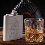 A stainless steel hip flask with an engraved design beside a glass of whiskey containing ice.