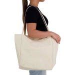 A beige tote bag with long handles, carried by a person, features a minimalist design and a logo.