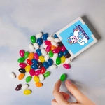 Assorted colorful jelly beans in various shades spilling from a 50g box. The box has a logo design.