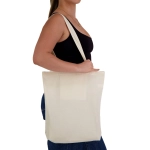 A cream-coloured tote bag is held by a person, featuring long straps and a simple design. It has a logo.