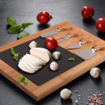 A bamboo cheese board featuring a black slate surface, sliced mozzarella, cherry tomatoes, and cheese knives.