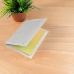 A sticky note pad with a grey cover and yellow sticky notes inside, placed on a wooden surface beside a potted plant.