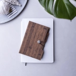 A wooden textured sticky note pad with an elastic closure, placed on top of a white notebook and a grey surface.