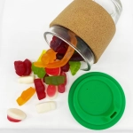 A glass coffee cup with a cork band, filled with colourful mixed lollies, and a green silicone lid.