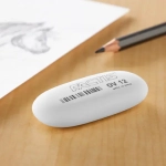 A white rubber eraser on a wooden surface beside a drawing and a pencil. The eraser features a logo.