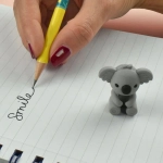 A grey koala-shaped pencil-top eraser next to a yellow pencil, with the word "Smile" written on lined paper.