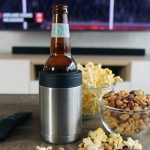 A stainless steel stubby holder in silver with a black silicone base, holding a bottle, surrounded by snacks.