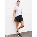 A model stands against a white wall wearing black sports shorts and a light grey t-shirt, paired with black sneakers.