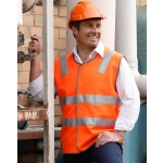 An orange hi-vis safety vest with reflective tapes, worn by a smiling man in a hard hat, stands near industrial equipment.