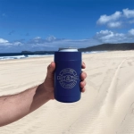 A drink bottle in a navy stubby holder, featuring a logo, on a sandy beach with a blue sky background.