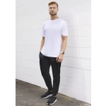 A man stands against a white wall wearing a plain white t-shirt and black track pants, with a watch and sneakers.