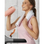A woman in a pink top holds a clear drink bottle while wearing a white towel around her neck, set in a gym environment.