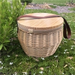 A round wicker picnic cooler basket with a wooden lid and a leather strap, set on grass with white flowers.