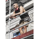 A man in a black sleeveless tee and shorts stands on scaffolding, wearing headphones and a tool belt.