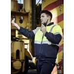A man in a hi-vis yellow and navy quilted jacket uses a walkie-talkie against an industrial background.