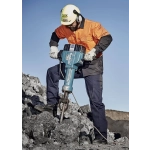 A man in an orange and black jacket and ear protection operates a blue jackhammer on rocky terrain.