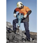 A man in safety gear operates a jackhammer on a rocky surface, wearing an orange shirt and navy blue cargo pants.
