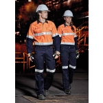 Two individuals wearing high-visibility orange shirts and navy cargo pants walk in an industrial setting, both wearing helmets and gloves.
