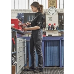 A woman in black rugged cargo pants stands at a workshop bench, engaging with tools and equipment. The pants feature pockets and a logo.