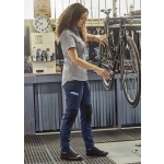 A woman in grey shirt and navy pants adjusts a bicycle in a workshop. The pants feature black knee patches and a logo.