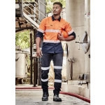 A man in orange and navy workwear with reflective stripes, holding a hard hat, stands in an industrial setting.