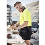 A man wearing a high-visibility yellow polo shirt and black rugby shorts measures wood on a construction site.