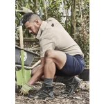 A man in a beige shirt and navy shorts kneels in a garden, working with a green shovel and wearing gloves and boots.