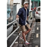 A man in a navy shirt and khaki shorts stands by a railing, with a skateboard and cars in the background.