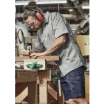 A man wears a grey short sleeve shirt with a logo, using a pencil at a workbench in a workshop setting.
