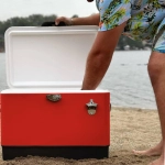 A metal ice cooler box in red, with a white lid, features a stainless steel latch and is placed on sandy beach terrain.