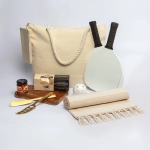 A cream tote bag with two paddles, a ball, a picnic blanket, a wooden board, knife, and snacks, arranged on a white background.