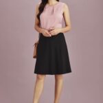 A woman stands wearing a pink top and a black flared skirt, accessorised with a handbag and high heels.